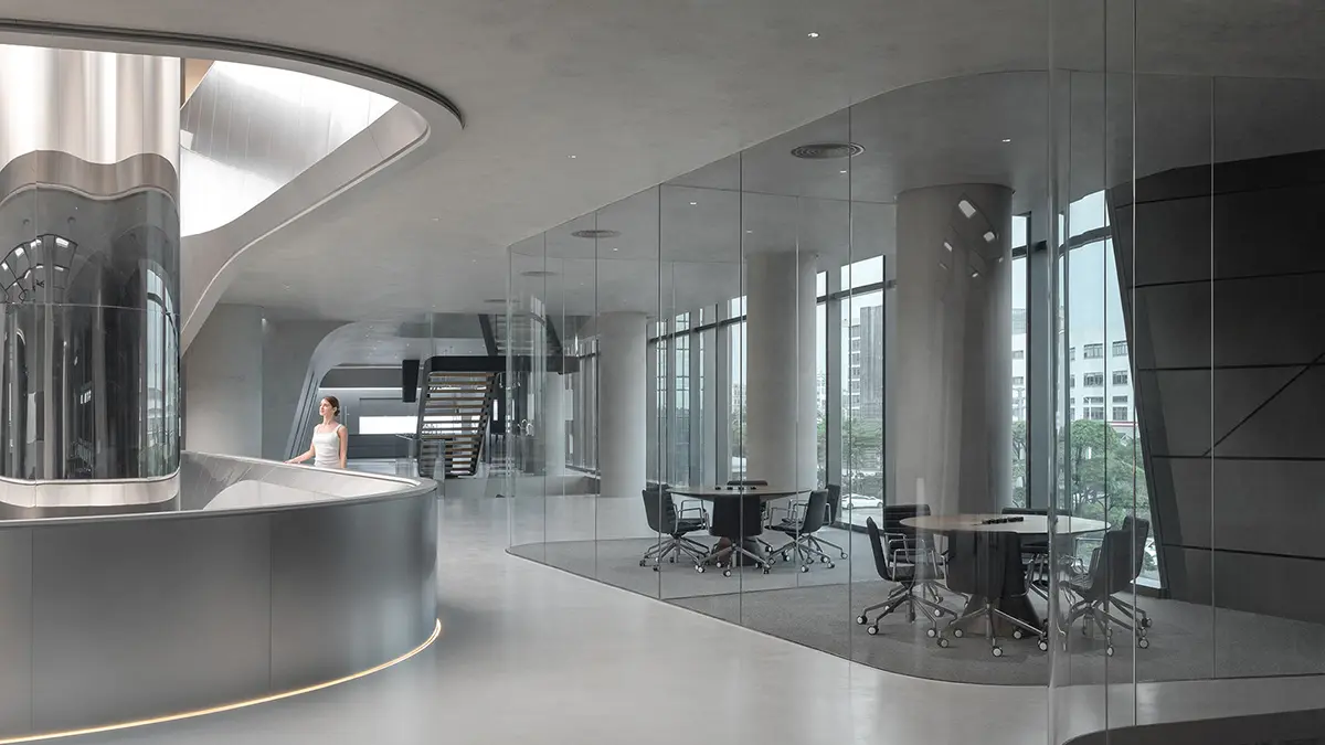 Futuristic office interior with curved metallic reception desk, glass-walled meeting rooms, round tables, and tall cylindrical columns. Natural light streams through large windows, highlighting the minimalist, open design and urban view outside.