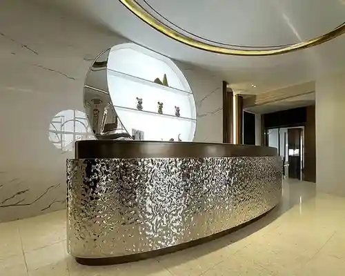 Elegant lobby reception area with a curved metallic desk, circular decorative panel, marble walls, and polished floor. A recessed ceiling with gold trim enhances the luxurious, modern atmosphere.